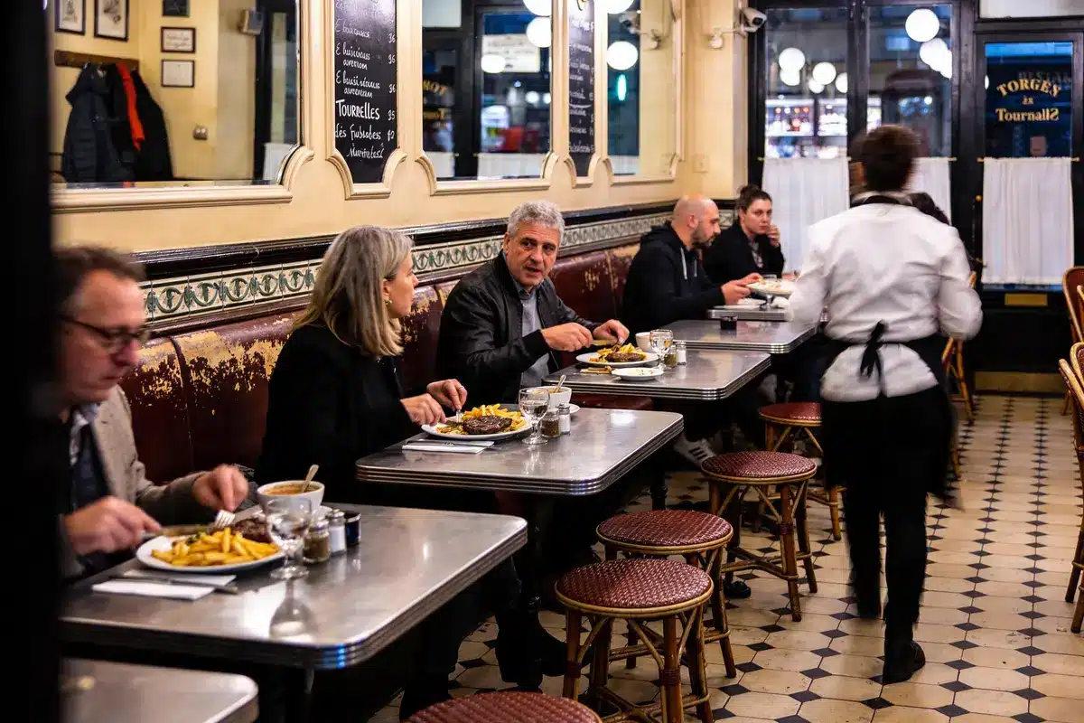 Des personnes dînent dans un bistrot parisien, assises à des tables en métal, plancher en damier.