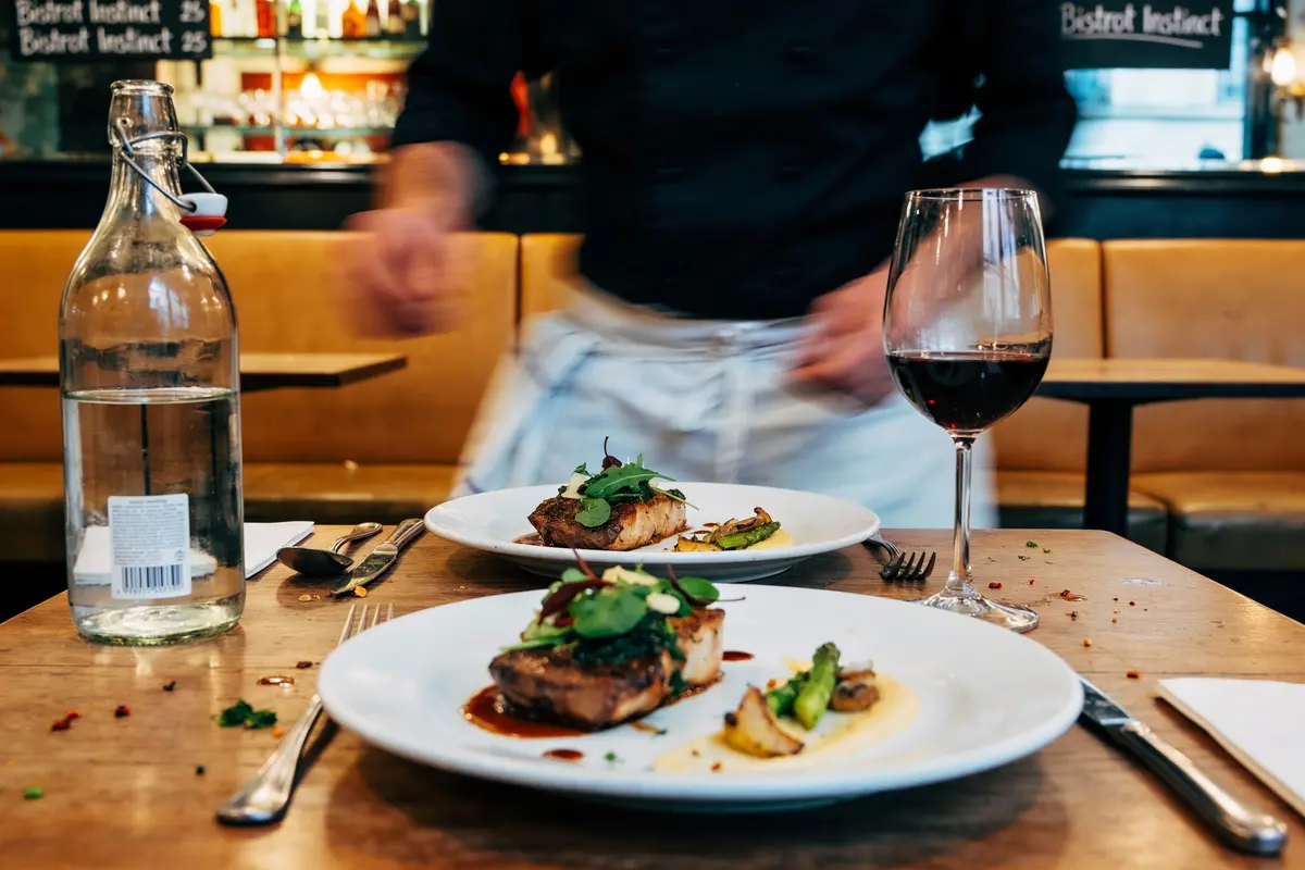 Deux assiettes gastronomiques, verre de vin rouge, bouteille d'eau, restaurant ambiance chaleureuse, serveur en mouvement.