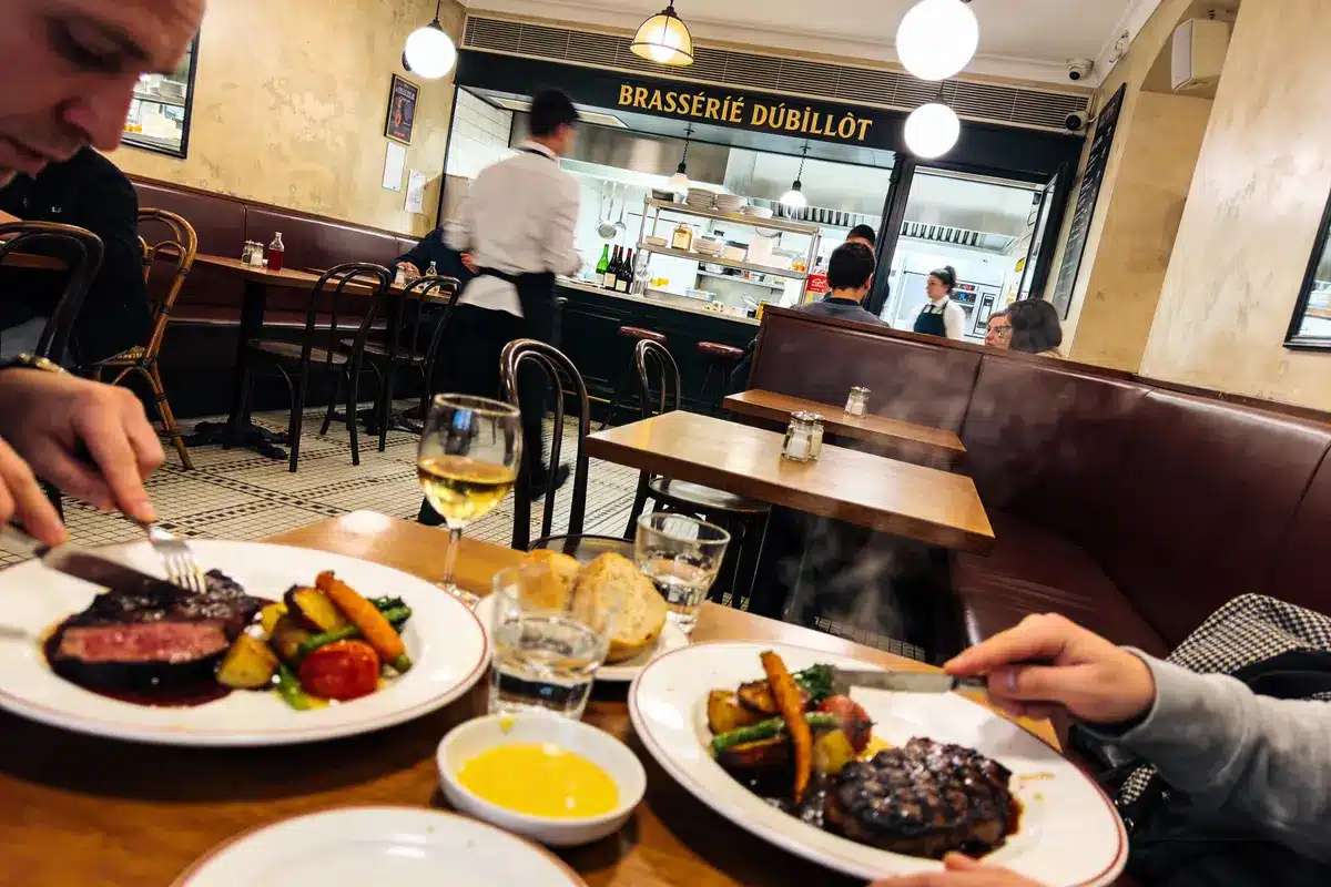 Repas dans une brasserie parisienne, deux assiettes de viande, légumes et vin, intérieur chaleureux et animé.
