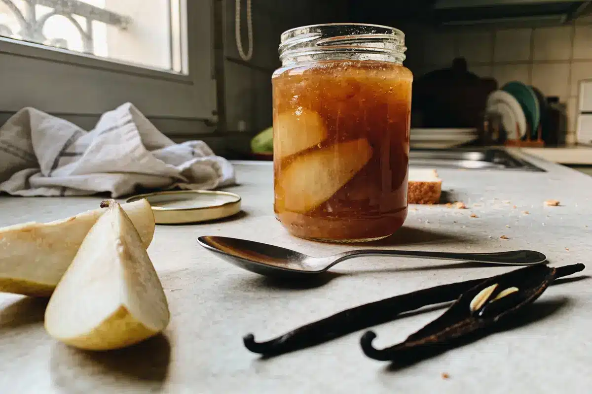 Bocal de confiture maison avec poires, vanille, cuillère, sur comptoir de cuisine ensoleillé.