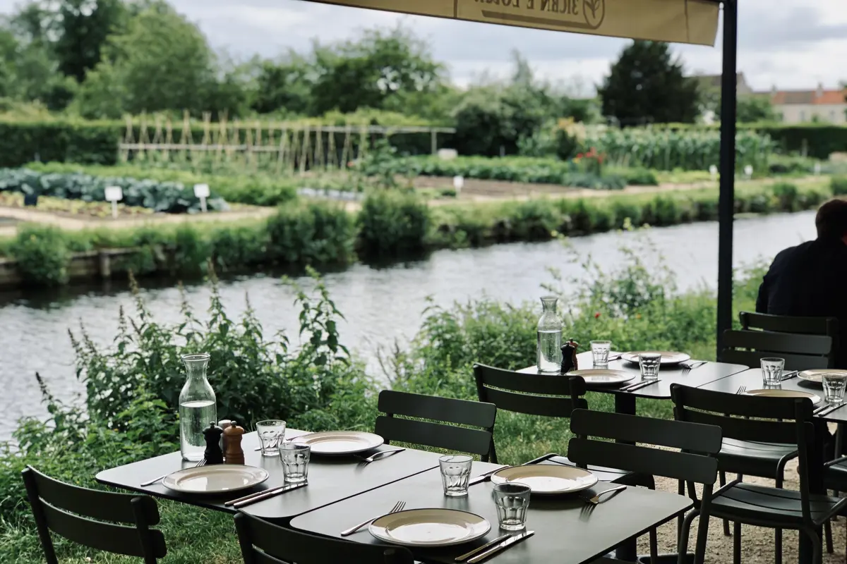 Terrasse de restaurant avec tables dressées, vue sur un jardin verdoyant et rivière en arrière-plan.
